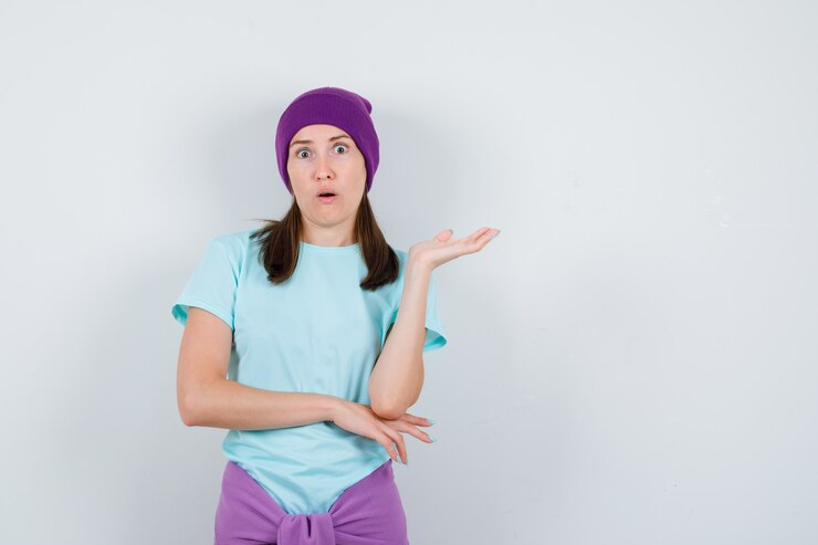 Foto mujer joven con la palma hacia fuera, manteniendo la boca abierta en camiseta azul, gorro morado y mirando sorprendido, vista frontal.