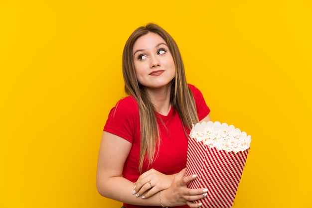 Mujer joven con palomitas de maíz sobre la pared rosada haciendo gestos de dudas mientras levanta los hombros