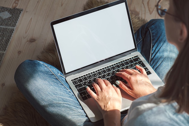 Mujer joven con pantalla en blanco de la computadora portátil para trabajar, sentada en el suelo en casa. Horarios flexibles y trabajo a distancia.