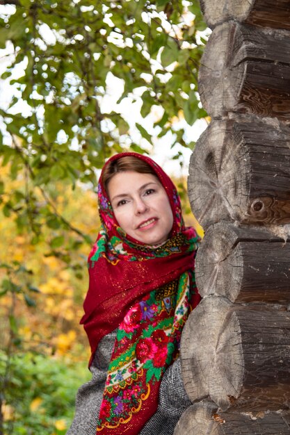 Foto una mujer joven con un pañuelo tradicional ruso mira a la vuelta de la esquina de una casa de madera. pavlovsk pañuelo rojo yaroslavl rusia 10.10.2019