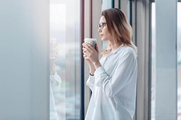 Mujer joven parada en la ventana de una oficina moderna y sosteniendo una taza de café