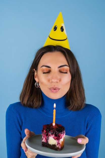 Mujer joven en una pared azul celebra un cumpleaños, sostiene un trozo de tarta, feliz, emocionado, pide un deseo, apaga la vela