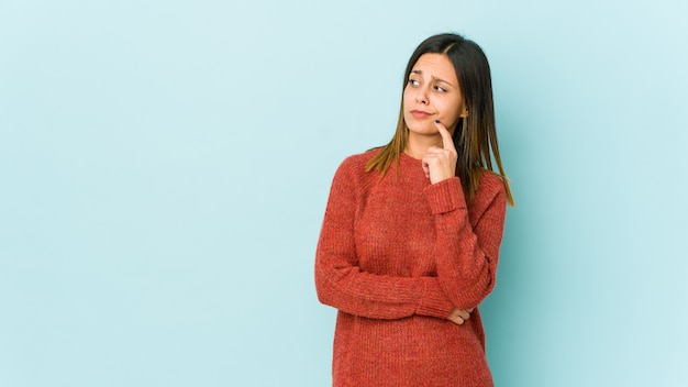 Mujer joven en la pared azul mirando hacia los lados con expresión dudosa y escéptica.