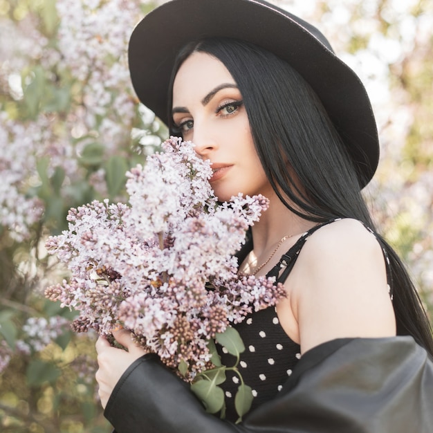 Mujer joven en el parque de flores descansa en la naturaleza. Chica de moda con sombrero elegante en vestido negro de moda y flores de primavera lila al aire libre. Hermosa dama huele increíble ramo de flores. Soleado retrato fresco