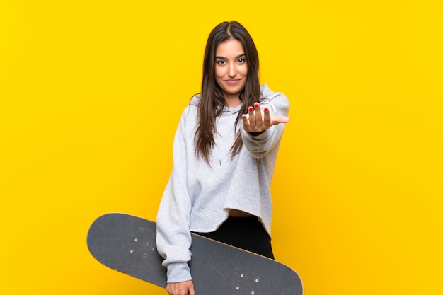 Mujer joven del patinador sobre la pared amarilla aislada que invita a venir con la mano. Feliz de que hayas venido