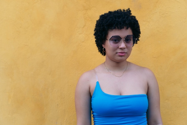 Mujer joven con peinado afro y gafas de sol mirando seriamente a la cámara con una blusa azul.