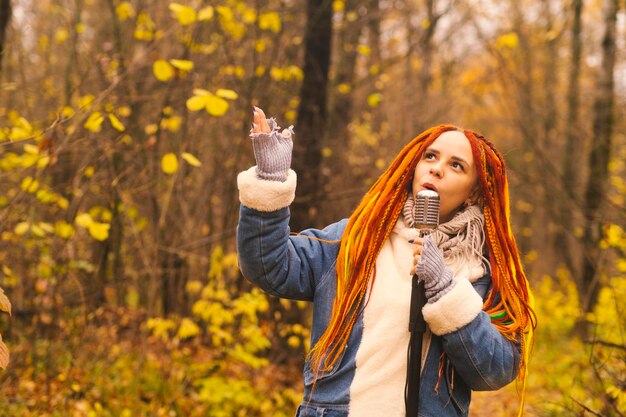 Mujer joven con peinado brillante con micrófono retro en el bosque de otoño Retrato de cantante femenina con rastas cantando en el micrófono en la naturaleza