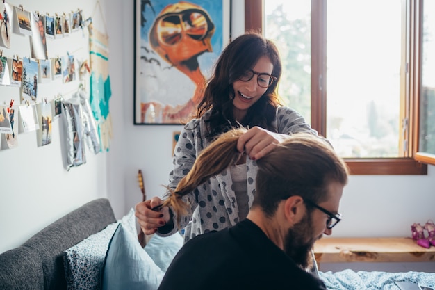 Mujer joven peinando el cabello del hombre barbudo interior casa dormitorio