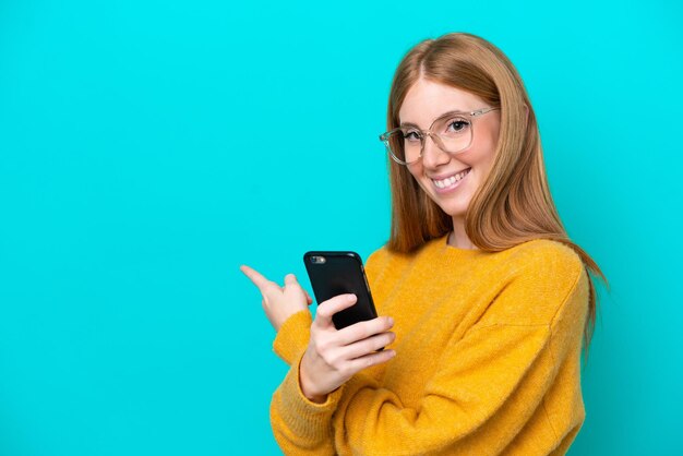 Mujer joven pelirroja aislada de fondo azul usando teléfono móvil y apuntando hacia atrás