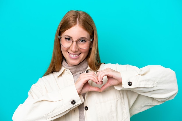 Mujer joven pelirroja aislada sobre fondo azul con gafas haciendo corazón con las manos