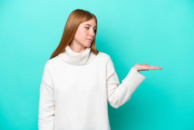 Mujer joven pelirroja aislada sobre fondo azul sosteniendo copyspace con dudas