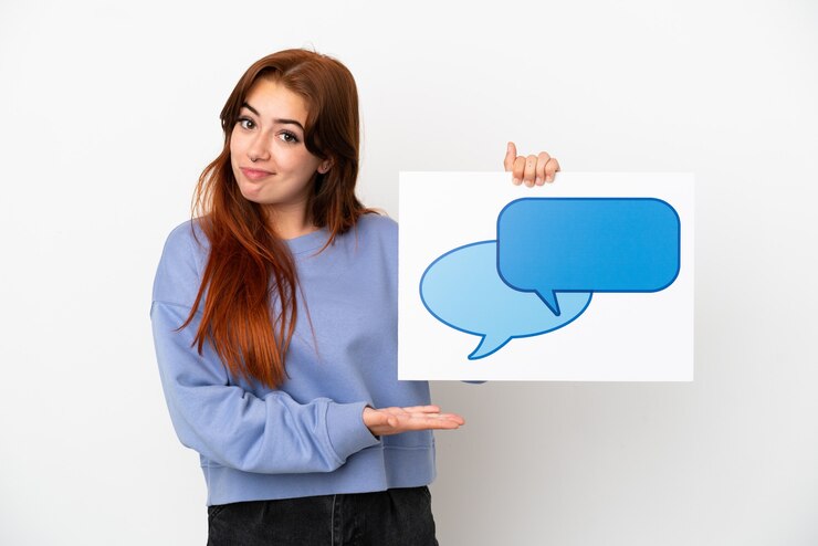Foto mujer joven pelirroja aislada sobre fondo blanco sosteniendo un cartel con el icono de la burbuja del discurso y apuntando hacia él