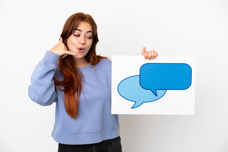 Foto mujer joven pelirroja aislada sobre fondo blanco sosteniendo un cartel con el icono de la burbuja del discurso y haciendo gesto de teléfono