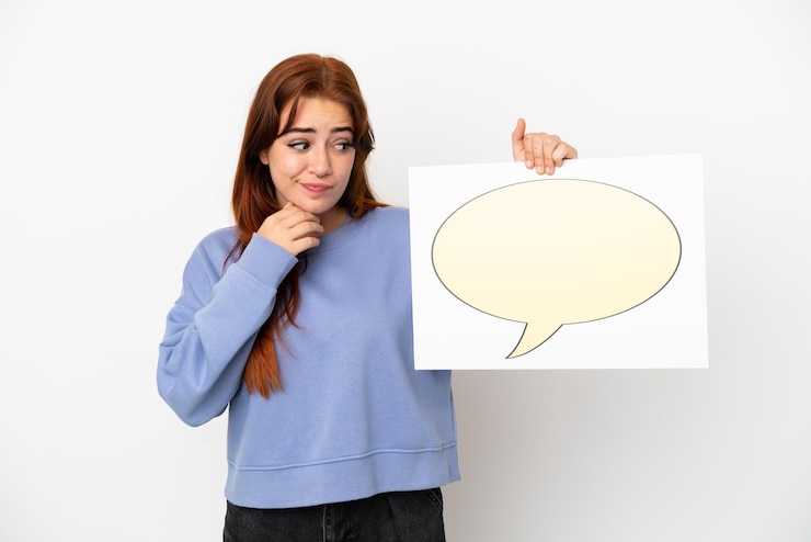 Foto mujer joven pelirroja aislada sobre fondo blanco sosteniendo un cartel con el icono de la burbuja del discurso y el pensamiento