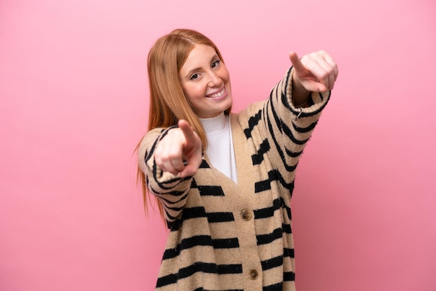 Mujer joven pelirroja aislada sobre fondo rosa apuntando al frente con expresión feliz