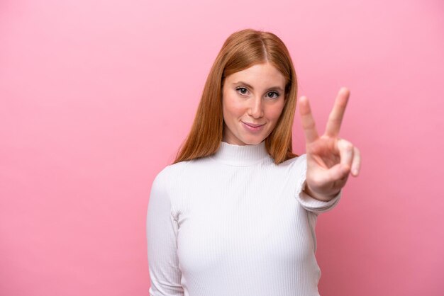 Mujer joven pelirroja aislada sobre fondo rosa sonriendo y mostrando el signo de la victoria