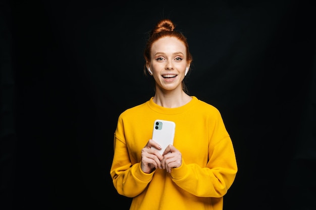 Mujer joven pelirroja alegre con auriculares inalámbricos sosteniendo el teléfono y escuchando música en línea en un fondo negro aislado Modelo de mujer pelirroja bonita que muestra emocionalmente expresiones faciales