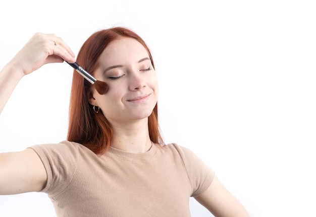 Mujer joven pelirroja alegre con pincel de maquillaje aislado sobre fondo blanco.