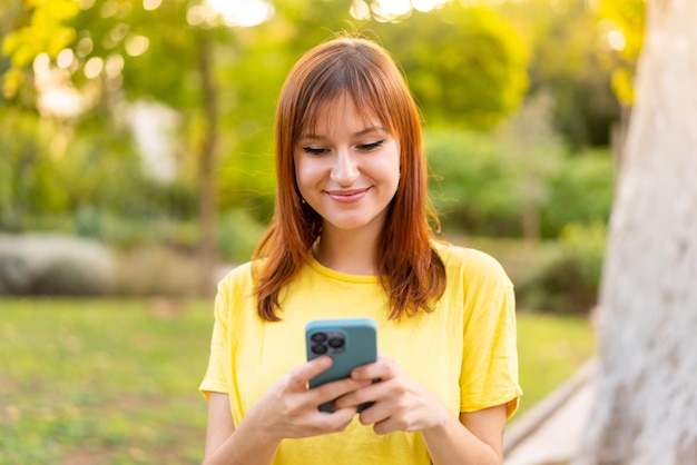 Mujer joven pelirroja bonita al aire libre enviando un mensaje o correo electrónico con el móvil