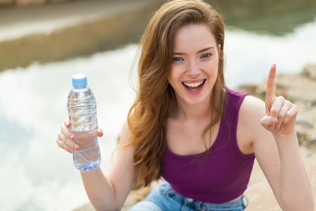 Foto mujer joven pelirroja con una botella de agua al aire libre señalando una gran idea