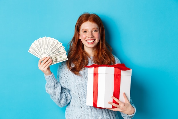 Mujer joven pelirroja con caja de regalo de Navidad y dinero, sonriendo complacido, de pie sobre fondo azul.