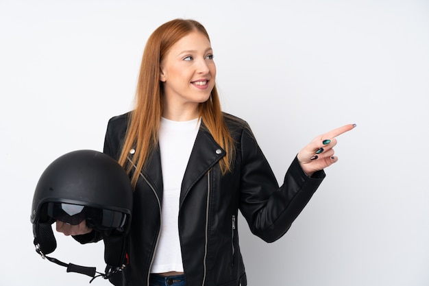 Mujer joven pelirroja con un casco de moto sobre pared blanca aislada apuntando hacia un lado para presentar un producto