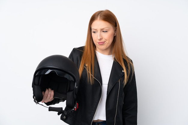 Mujer joven pelirroja con un casco de moto sobre pared blanca aislada con expresión triste