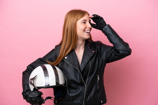 Mujer joven pelirroja con un casco de motocicleta aislado sobre fondo rosa sonriendo mucho