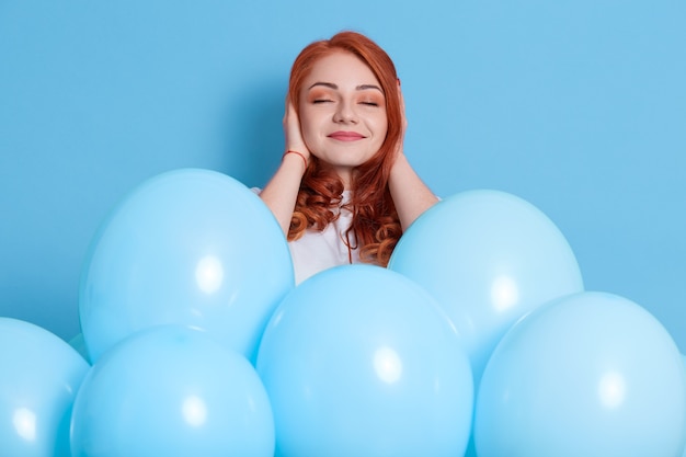 Mujer joven pelirroja complacida en ropa casual blanca que cubre las orejas con los dedos aislados en la pared de color, posando entre globos de aire, celebración, sorpresa en la fiesta, chica con sonrisa y ojos cerrados