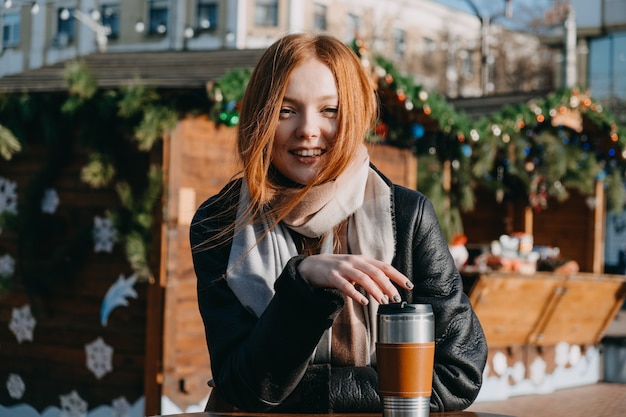 Mujer joven pelirroja disfrutando del sol y bebiendo café para llevar vagando por las calles de la ciudad de invierno