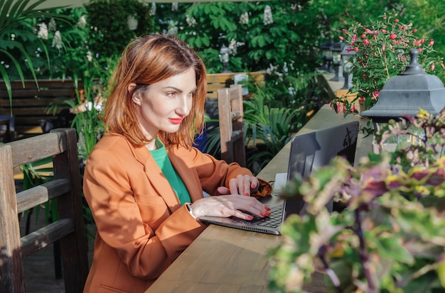 Mujer joven pelirroja con una elegante chaqueta se sienta en la terraza abierta de un café con una computadora portátil. Trabajar fuera de la oficina, autónomo, prepararse para una reunión de negocios