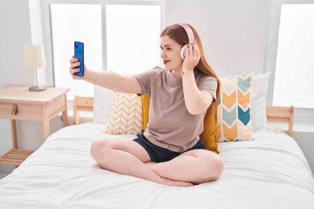 Mujer joven pelirroja escuchando música hacer selfie por teléfono inteligente en el dormitorio