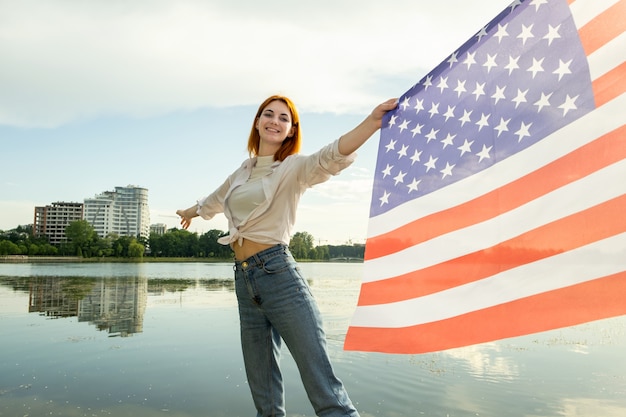 Mujer joven pelirroja feliz con la bandera nacional de Estados Unidos en su mano. Chica positiva celebrando el día de la independencia de Estados Unidos.