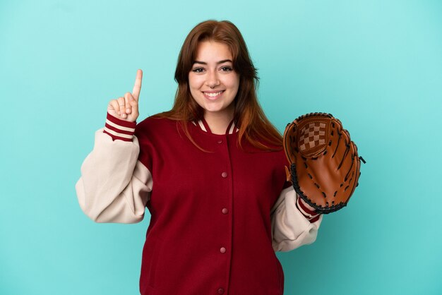 Mujer joven pelirroja jugando béisbol aislado sobre fondo azul apuntando hacia una gran idea