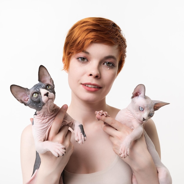 Mujer joven pelirroja mirando a la cámara abrazando a dos Sphynx Cat en manos sobre fondo blanco.