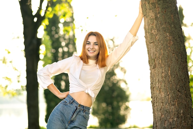 Mujer joven pelirroja positiva en blusa blanca y jeans de pie en el parque de verano disfrutando de un día cálido.