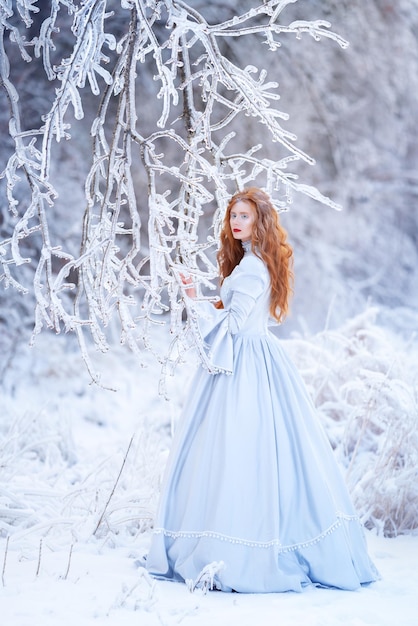 Mujer joven pelirroja una princesa camina en un bosque de invierno con un vestido azul Frost y nieve