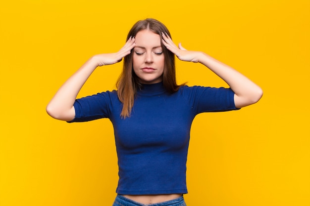 Mujer joven pelirroja que parece concentrada, reflexiva e inspirada, lluvia de ideas e imaginación con las manos en la frente en una pared plana