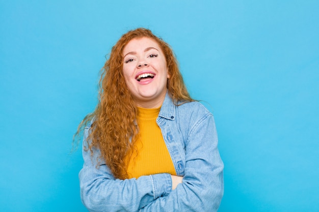 Foto mujer joven pelirroja que parece un triunfador feliz, orgulloso y satisfecho sonriendo con los brazos cruzados en la pared azul