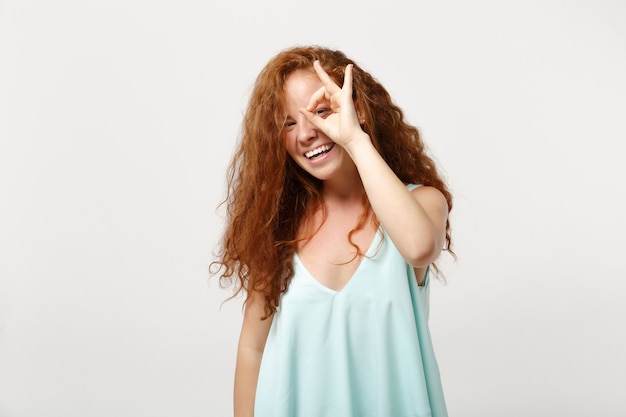 Mujer joven pelirroja riendo en ropa ligera casual posando aislada sobre fondo de pared blanca. Concepto de estilo de vida de personas. Simulacros de espacio de copia. Sosteniendo la mano cerca de los ojos, imitando anteojos o binoculares.