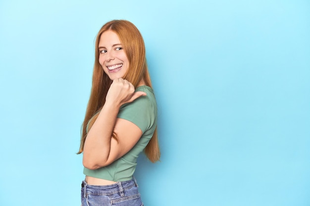 Mujer joven pelirroja sobre fondo azul señala con el dedo pulgar lejos riendo y despreocupada