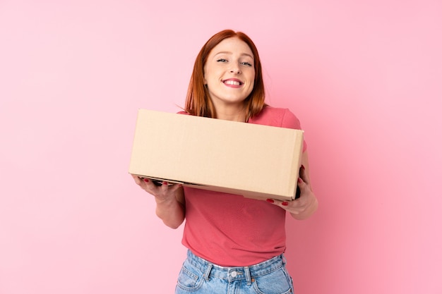 Mujer joven pelirroja sobre fondo rosa aislado sosteniendo una caja para moverlo a otro sitio