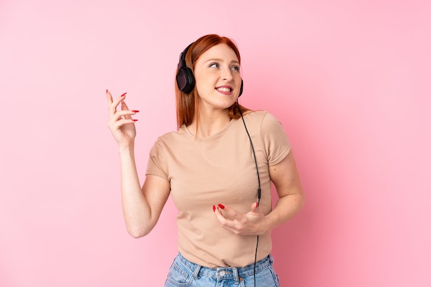 Mujer joven pelirroja sobre fondo rosa aislado usando el móvil con auriculares y baile