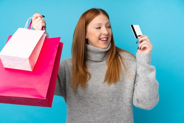 Mujer joven pelirroja sobre pared azul aislada con bolsas de compras y una tarjeta de crédito