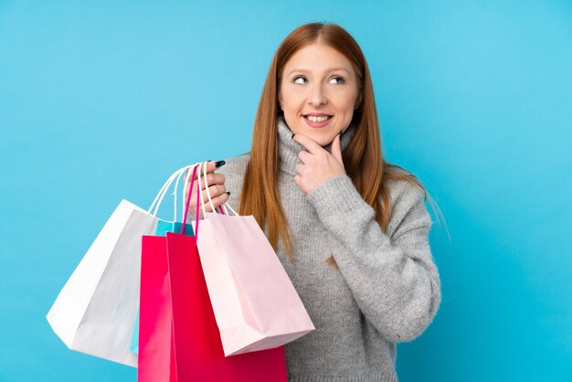 Mujer joven pelirroja sobre pared azul aislada sosteniendo bolsas de compras y pensando