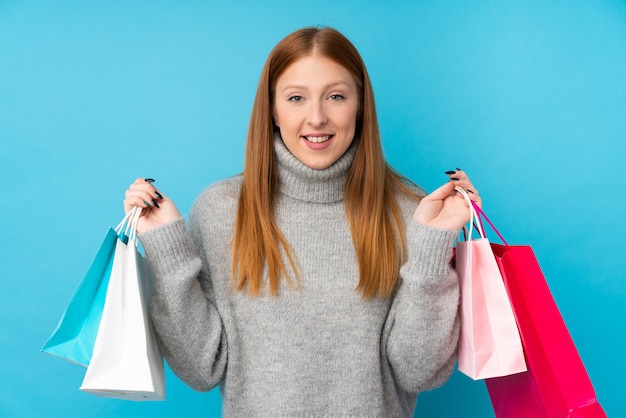 Mujer joven pelirroja sobre pared azul aislada sosteniendo bolsas de compras y sonriendo