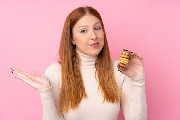 Mujer joven pelirroja sobre pared rosa aislado con coloridos macarons franceses y haciendo gesto de dudas