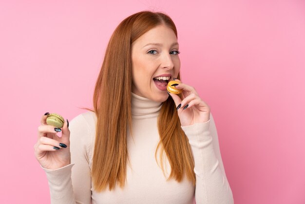 Mujer joven pelirroja sobre pared rosa aislado sosteniendo coloridos macarons franceses y comiéndolo