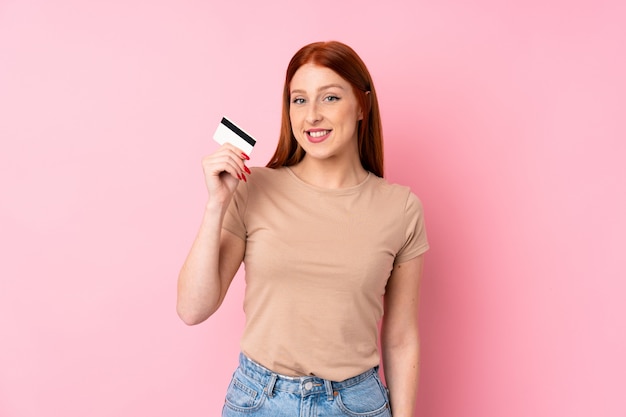 Mujer joven pelirroja sobre pared rosa aislado con una tarjeta de crédito