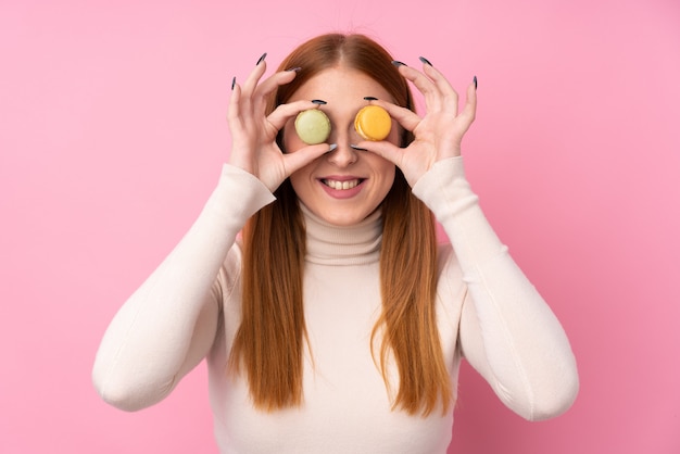 Mujer joven pelirroja sobre pared rosa aislado vistiendo coloridos macarons franceses como gafas